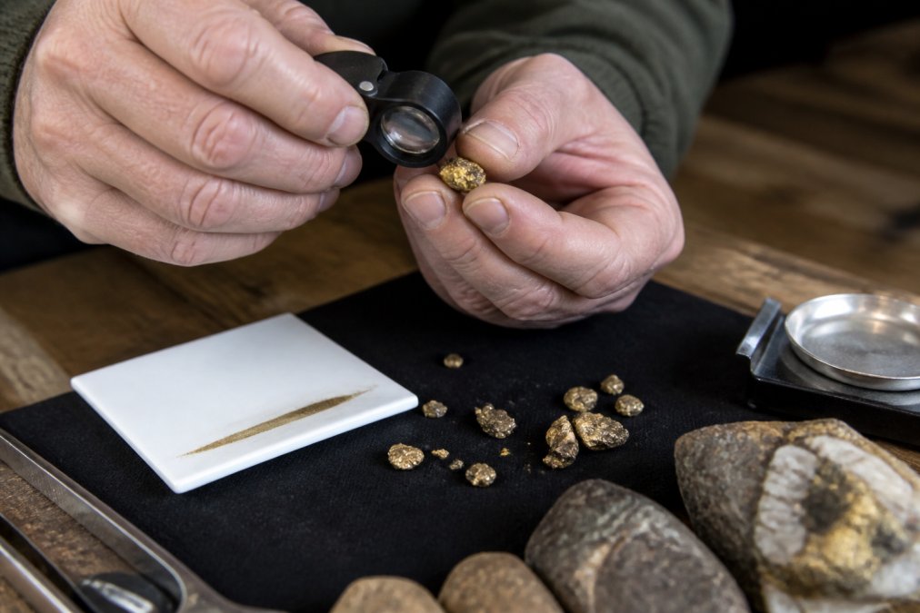 Person using a magnifying loupe to examine a raw gold specimen on a dark surface with a streak plate nearby
