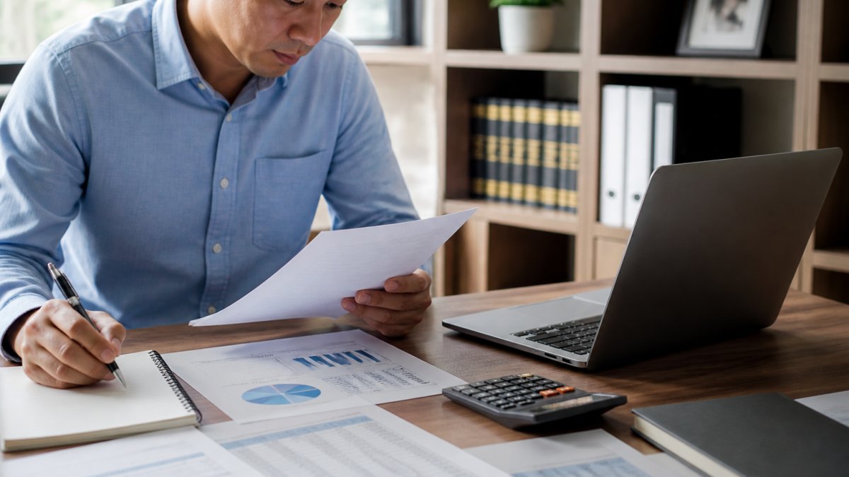 Professional reviewing financial documents and asset portfolio at desk with calculator, notepad, and laptop showing spreadsheets