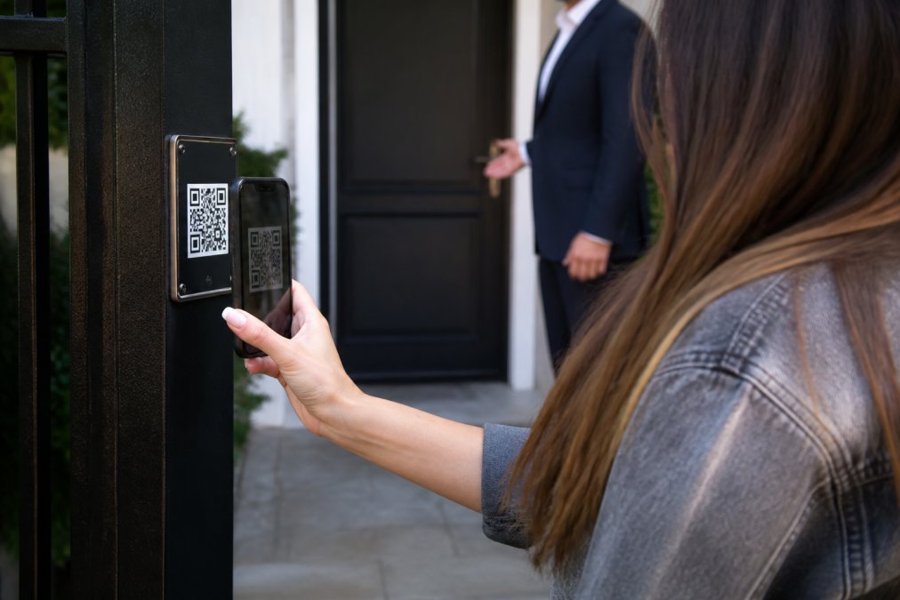 Close-up of smartphone screen displaying a QR code being scanned outside a residential property entrance, with a visitor's hand holding the phone and a property door visible in background