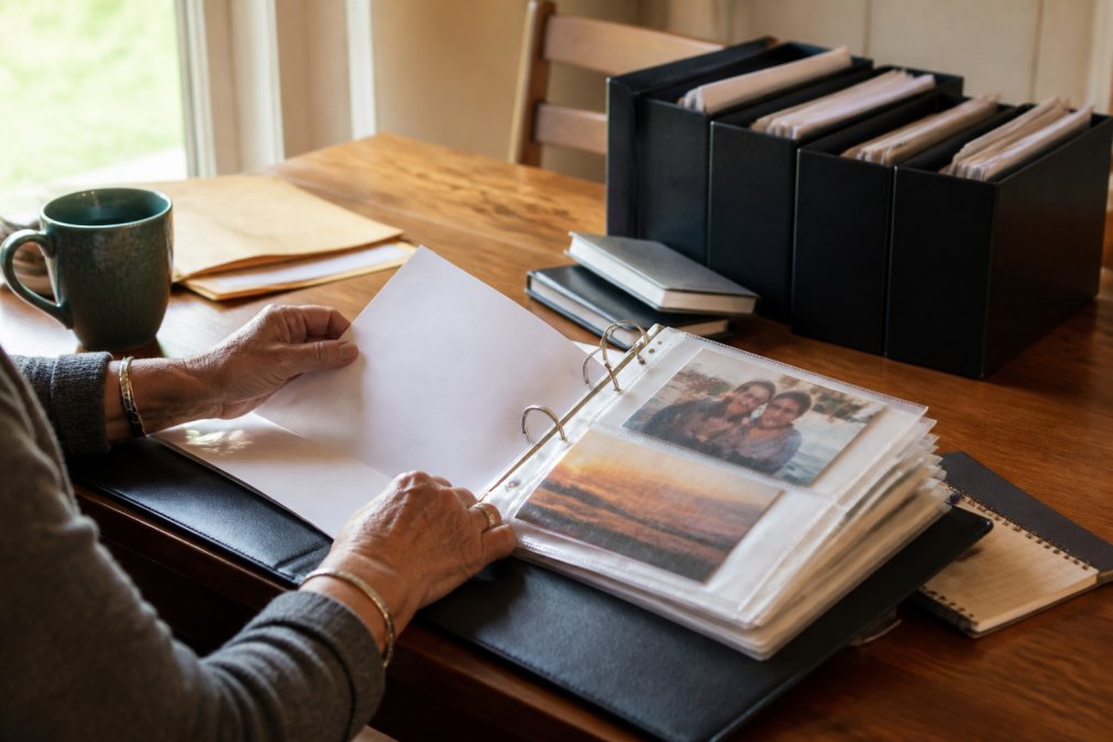 Hands carefully organizing important documents, photographs, and labeled folders into a well-organized binder on a wooden desk in natural light