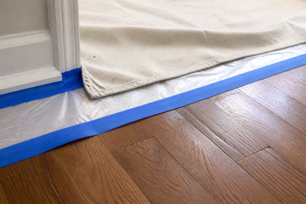 Close-up of layered canvas and plastic drop cloths on hardwood flooring with painter's tape securing edges, showing proper floor protection technique with natural light highlighting the material textures