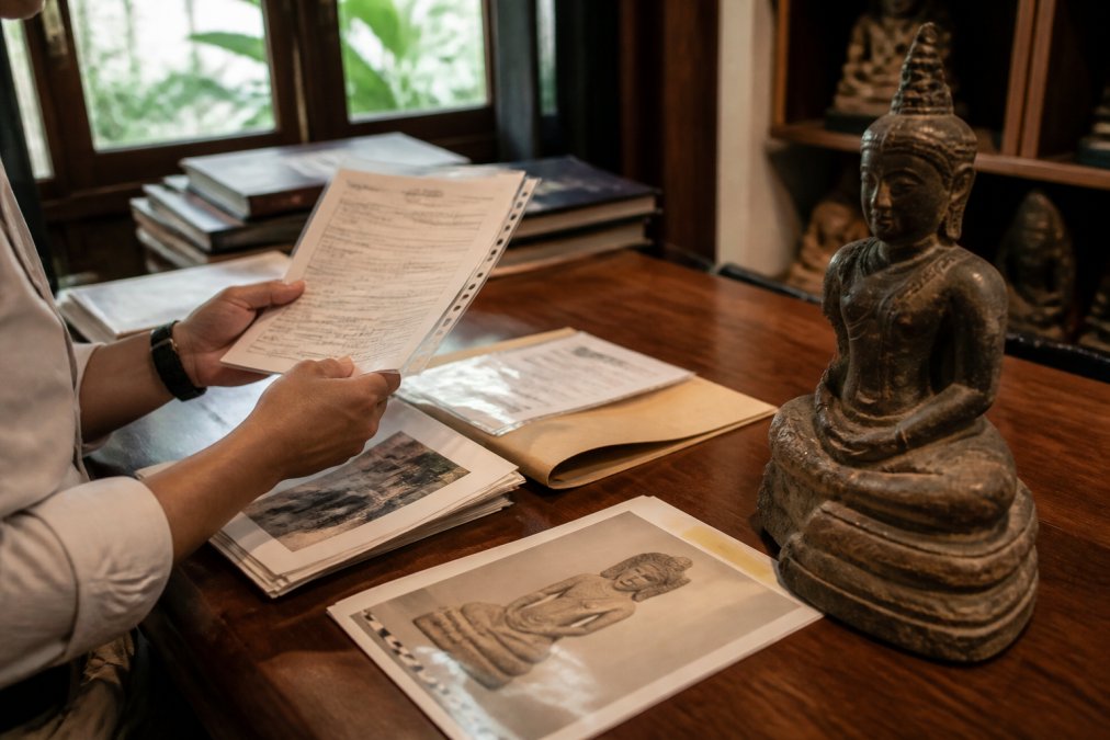 Manos de coleccionista examinando cuidadosamente permisos de exportación históricos, documentación arqueológica y documentos de procedencia junto a una escultura jemer expuesta en un escritorio de madera con iluminación natural en la ventana