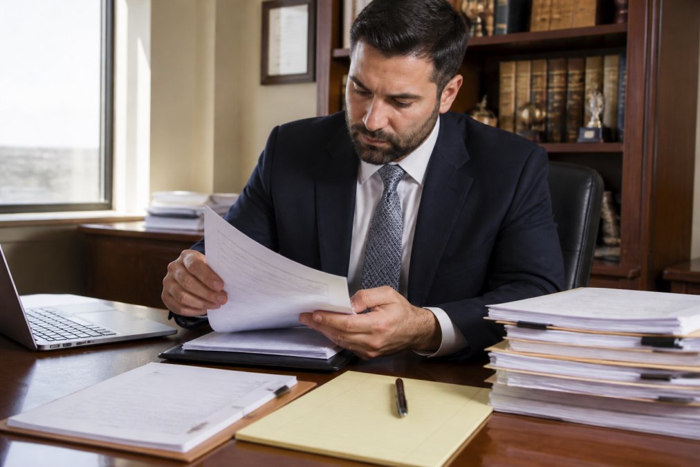 Professional reviewing case files and claim documents at desk with laptop, legal notepad, and stacked papers showing investigation work, focused lighting on documents