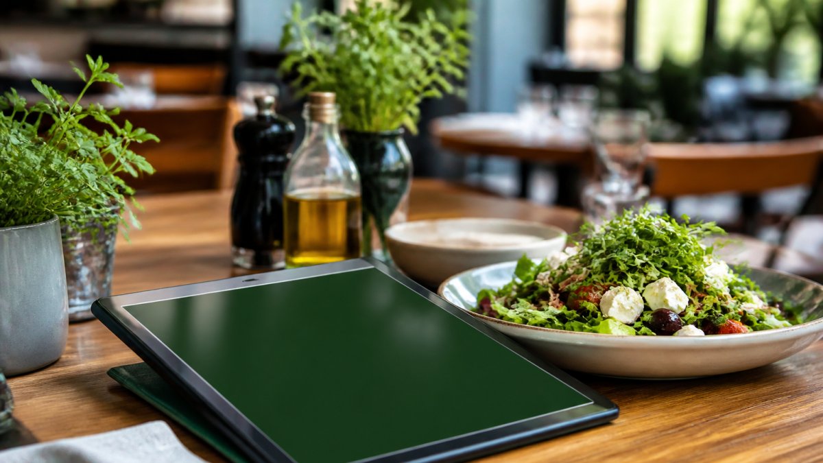 Primer plano de una tableta sobre una mesa de restaurante mostrando un menú digital con fondo verde y tipografía blanca, junto a un plato de ensalada fresca y hierbas, luz natural de mediodía