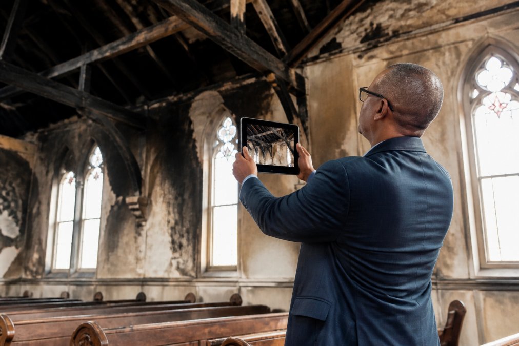 Church administrator photographing structural damage to sanctuary interior with tablet, documenting details of fire-damaged wooden beams and scorched walls under natural light from windows