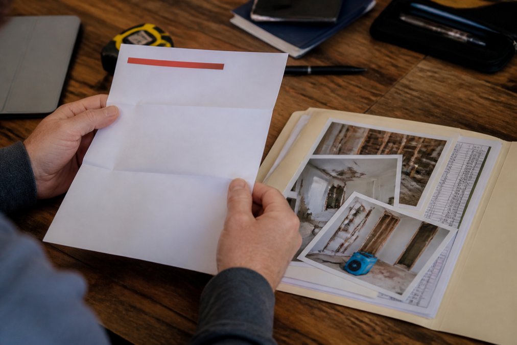 Close-up of hands holding denied insurance claim letter next to folder containing photographs of property damage and repair estimates on wooden desk