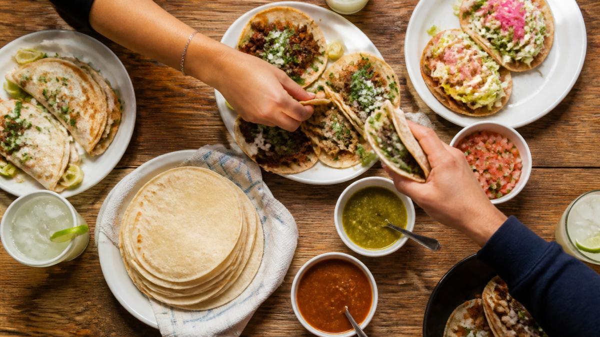 Overhead shot of a restaurant table crowded with tacos, a plate of handmade tortillas, margaritas, and small bowls of salsa, with two people reaching in to share the food