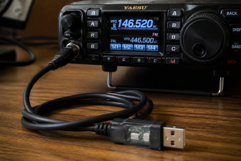 Close-up of a professional-grade USB programming cable with FTDI chipset connector attached to a Yaesu ham radio on a desk, showing the frequency dial and memory channel display in the background
