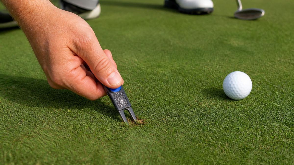 Close-up of a golfer's hand using a divot repair tool on a green after marking a ball, showing the tool in action and the repaired spot with proper spike aeration visible