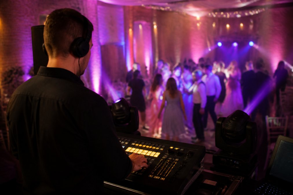 A mobile DJ at a wedding reception, hands on a lighting controller while colored uplights and moving head fixtures illuminate the dance floor behind them