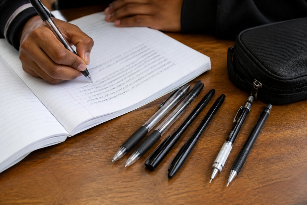 Close-up of a student's hand writing notes with a black ballpoint pen on a lined notebook, with gel pens and mechanical pencils arranged neatly beside the notebook on a wooden desk