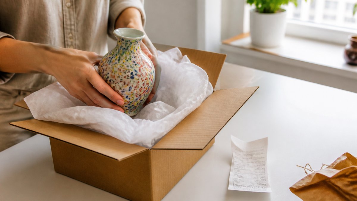 A person carefully repacking a colorful ceramic vase into its original box with tissue paper, a paper receipt resting on a clean white table beside it, soft daylight from a nearby window
