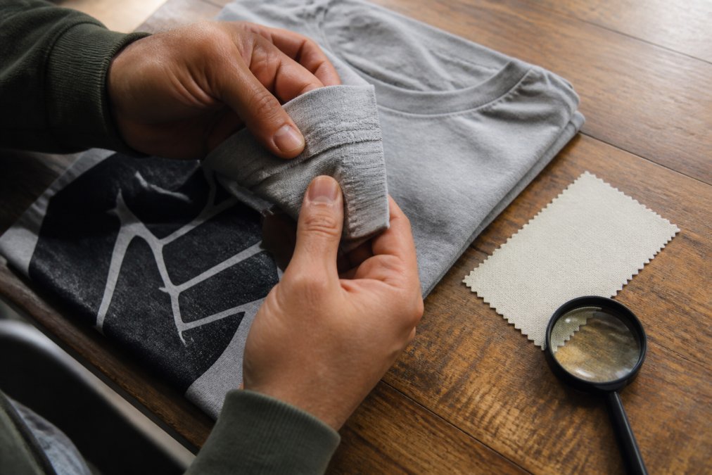Close-up of hands inspecting the stitching and printed graphic on a folded cotton t-shirt on a wooden table, with a fabric swatch and magnifying glass nearby