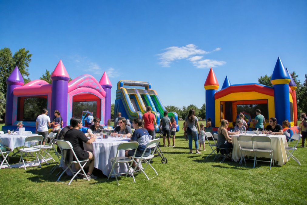 Professional event setup showing bounce houses, tables, and chairs arranged at an outdoor celebration with families enjoying the equipment under clear skies