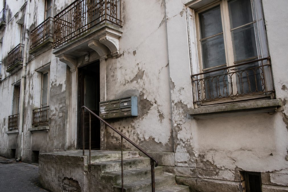 Façade d'immeuble collectif avec peinture écaillée par endroits, balcons en fer forgé montrant de la rouille visible, boîtes aux lettres endommagées ou cassées, escalier d'entrée aux marches usées et plinthe endommagée, fenêtres communes mal entretenues