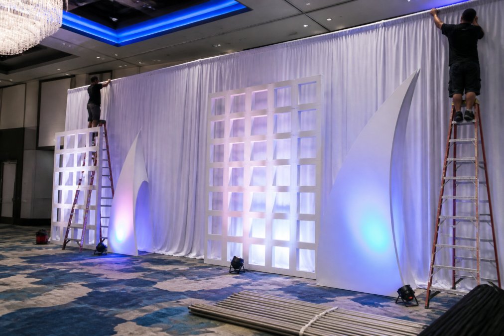 Professional installation crew setting up floor-to-ceiling pipe and drape backdrop with custom scenic elements and professional lighting at a modern corporate venue, workers on ladders securing drapes to ceiling grid