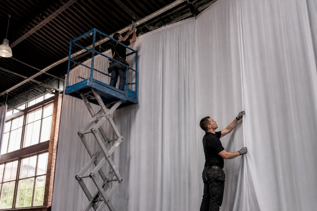 Professional installation crew securing pipe and drape framework to venue ceiling with rigging equipment, while another team member adjusts fabric tension on a 25-foot-tall drape panel in a modern event space with industrial architecture