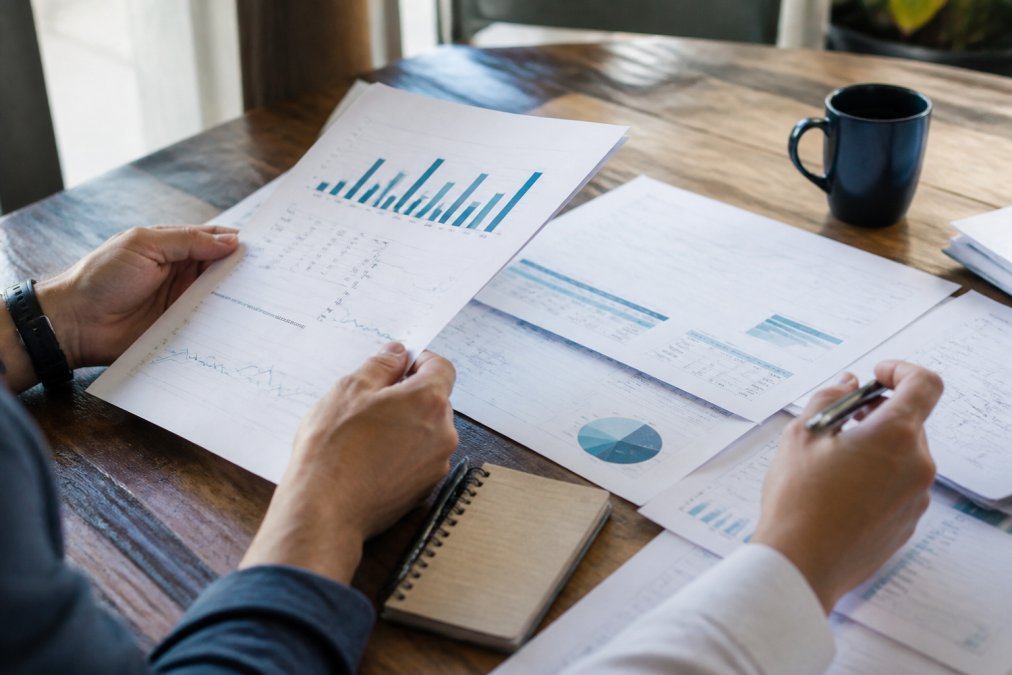 Close-up of hands holding real estate market analysis documents showing price comparisons, days on market statistics, and neighborhood appreciation data spread across table