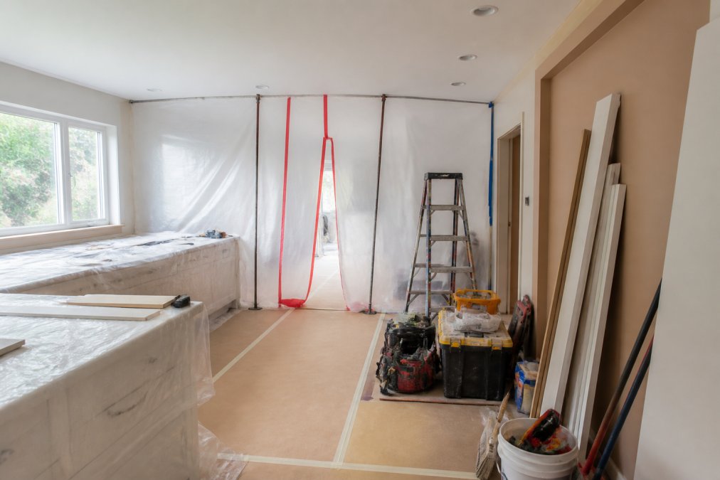 Kitchen actively under renovation showing dust containment barriers, work surfaces covered with protective plastic, organized tool staging areas, and construction materials neatly arranged in the workspace