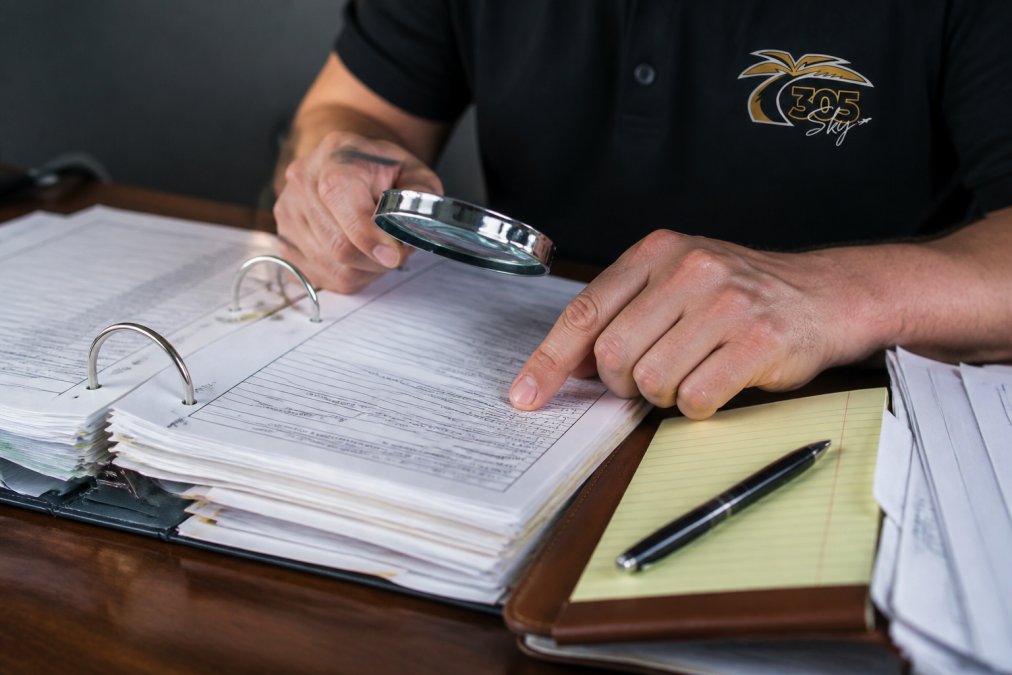 Close-up of hands reviewing aircraft logbooks and maintenance documentation at a desk with a magnifying glass and notes, natural office lighting