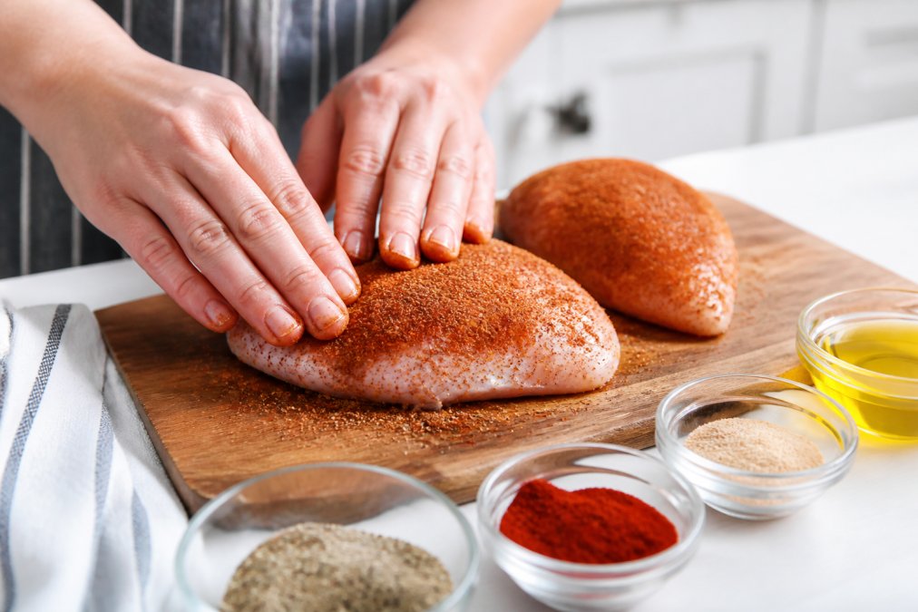 Hands pressing a dry rub seasoning blend onto raw chicken breasts on a wooden cutting board in a bright kitchen