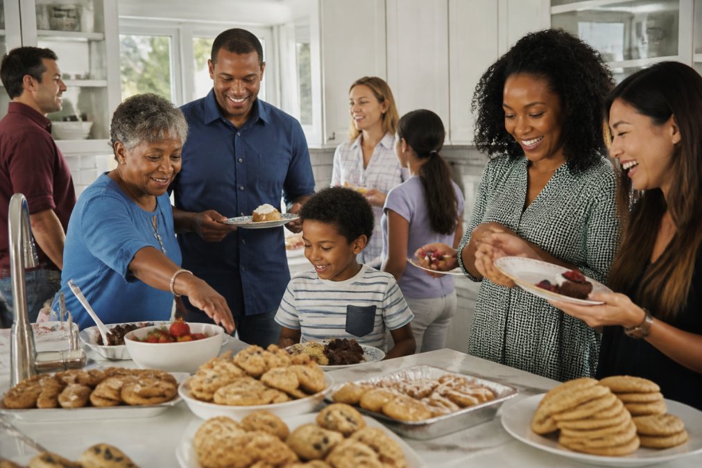 Diverse group of people of different ages, races, and genders enjoying fresh-baked cookies and teacakes in a warm home kitchen with natural afternoon light, genuine smiles, some holding plates of treats