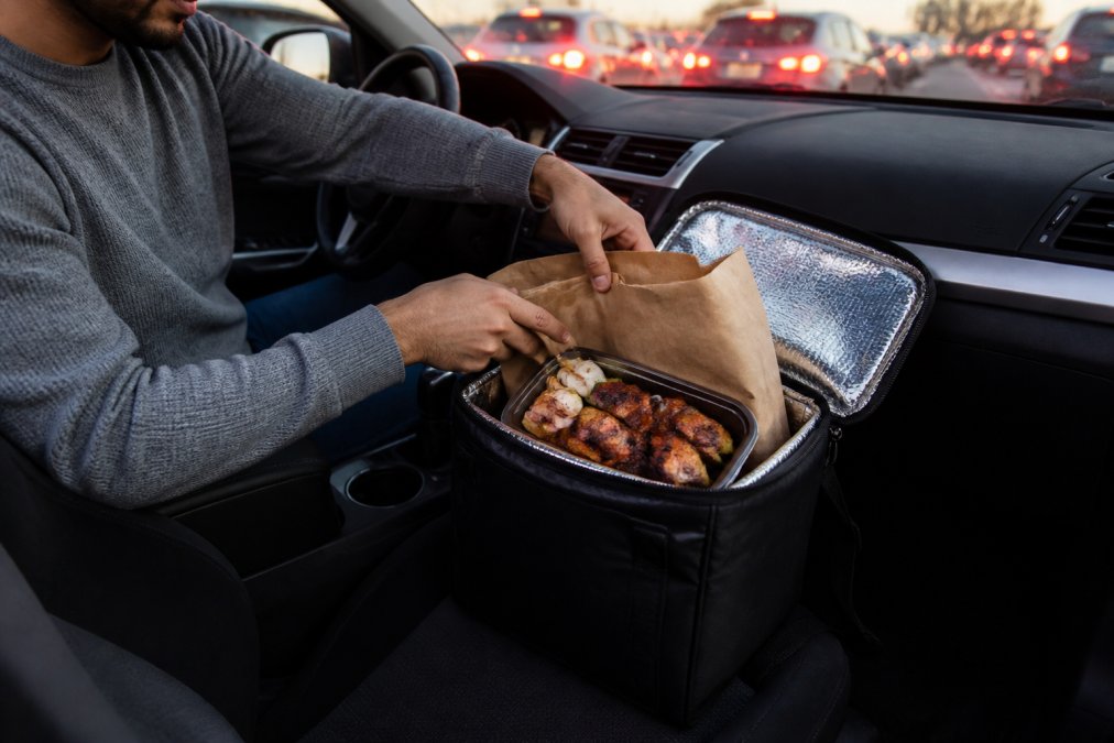 A commuter placing a takeout bag into an insulated cooler bag on the passenger seat of a car, with a warm meal visible inside, set during evening rush hour