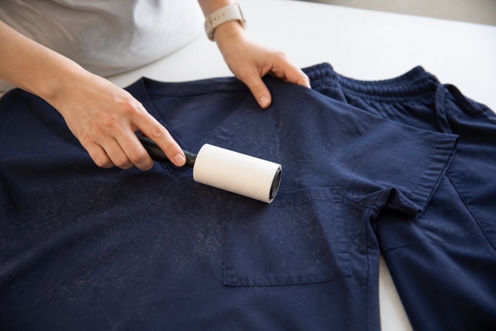 Close-up of hands using a lint roller on quick-drying scrub fabric with visible pet hair, demonstrating pre-wash preparation technique on a white surface with natural lighting