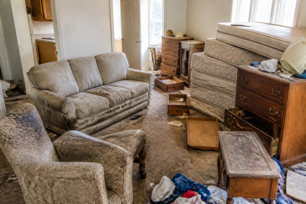 Cluttered rental unit interior with abandoned upholstered furniture, old wooden dressers, stacked mattresses, and scattered household items awaiting removal in natural daylight