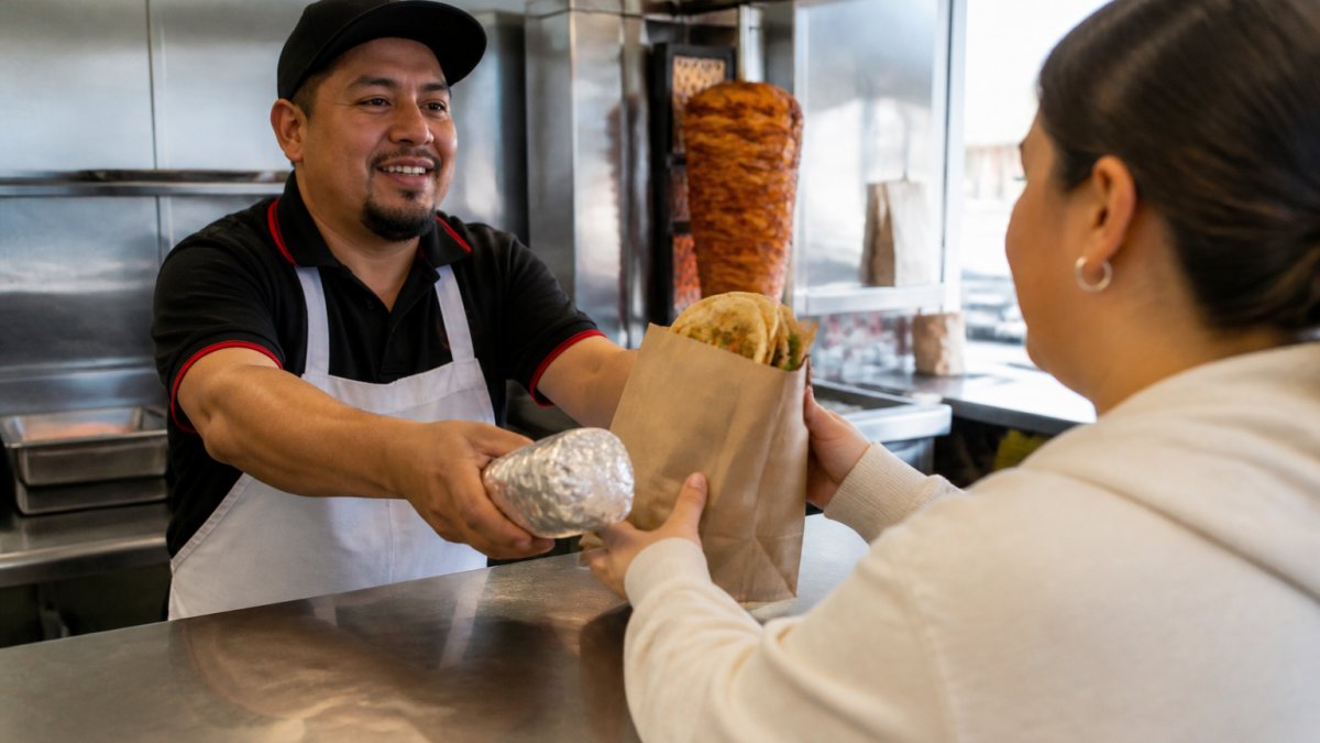 A counter server handing a wrapped burrito and a paper bag of tacos to a customer in a bright neighborhood taqueria, with a grill visible behind the counter