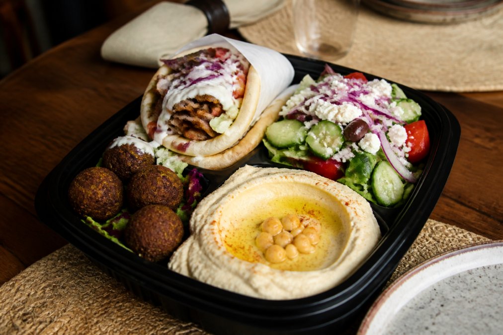 A delivery container with fresh Mediterranean food including gyro, hummus, falafel, and salad ready to be served at a dining table