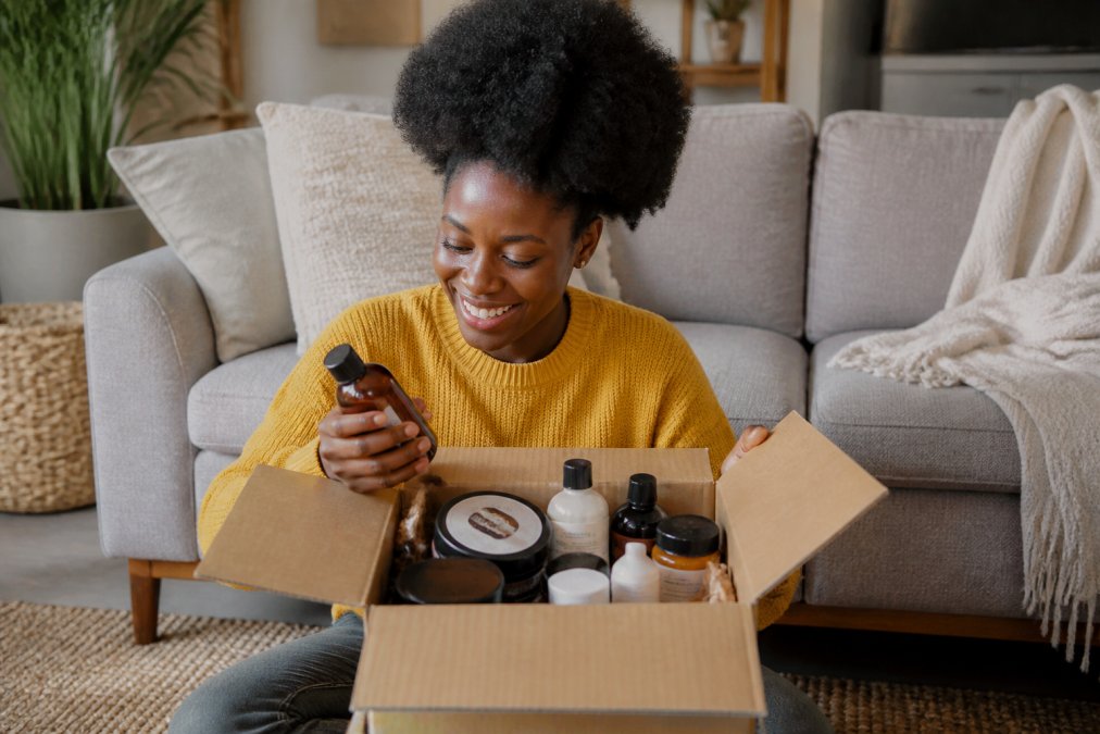 A woman with 4C hair smiling while opening a box of quick shipping beauty supply for natural hair at home.