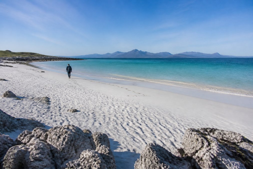 A solitary visitor walking along white coral sand beach with turquoise water and distant Outer Hebrides views on a clear morning, soft sunlight creating shadows across the rippled sand