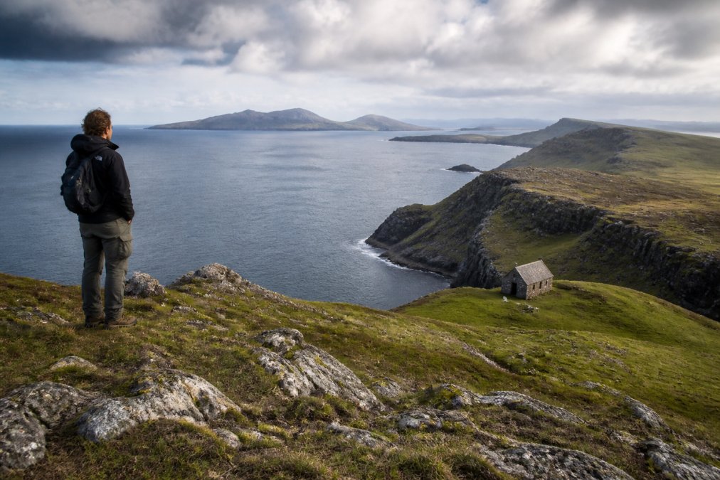 A hiker standing at a dramatic clifftop viewpoint overlooking the sea with a small stone bothy visible, expansive coastal landscape and distant islands in background, wind-swept grasses in foreground