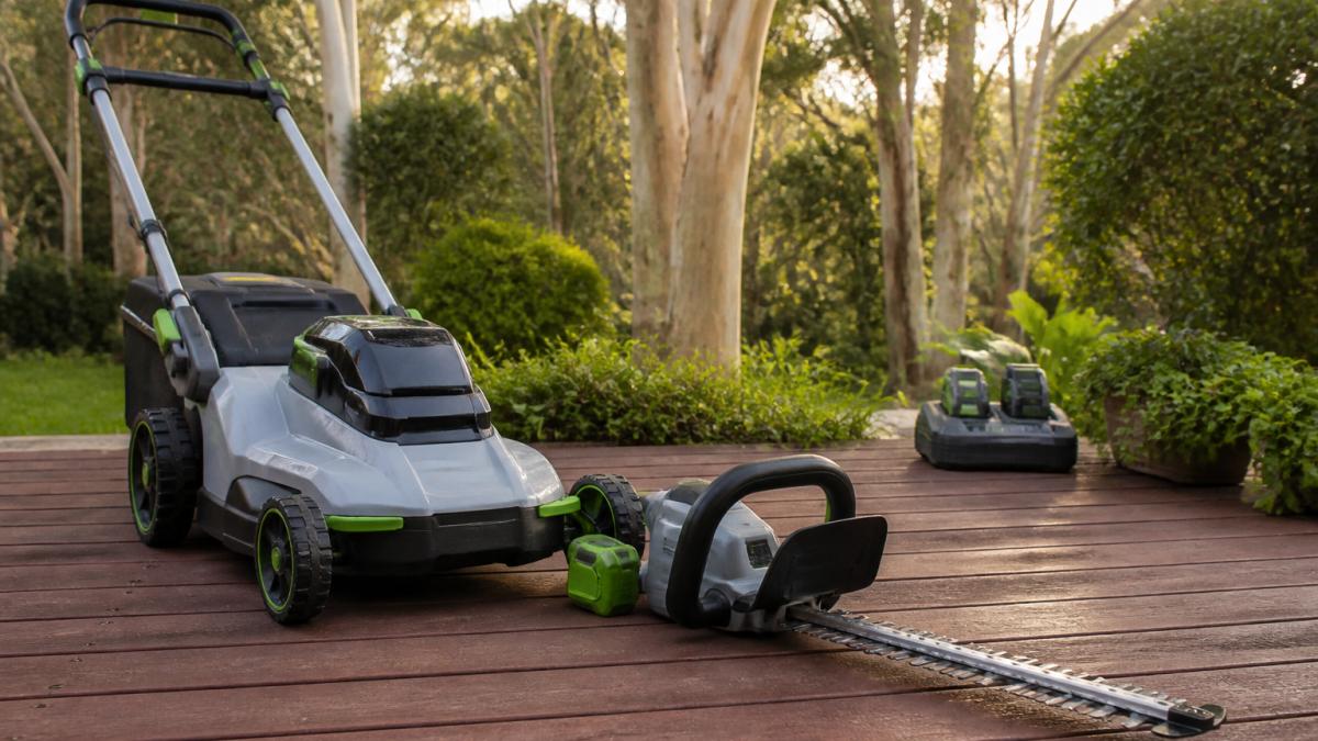 Close-up of a battery-powered lawn mower and hedge trimmer resting on a timber deck in a leafy garden, charging station visible in the background, soft morning light through gum trees