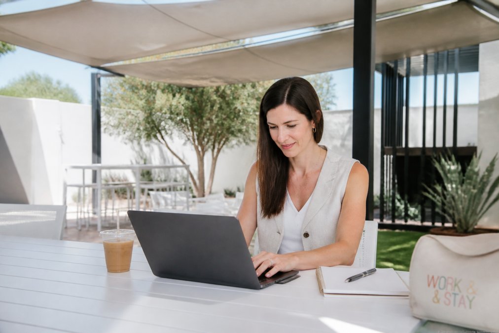 Professional parent working on laptop under shade structure in outdoor workspace with ergonomic desk setup, beverage, and notebook in natural daylight