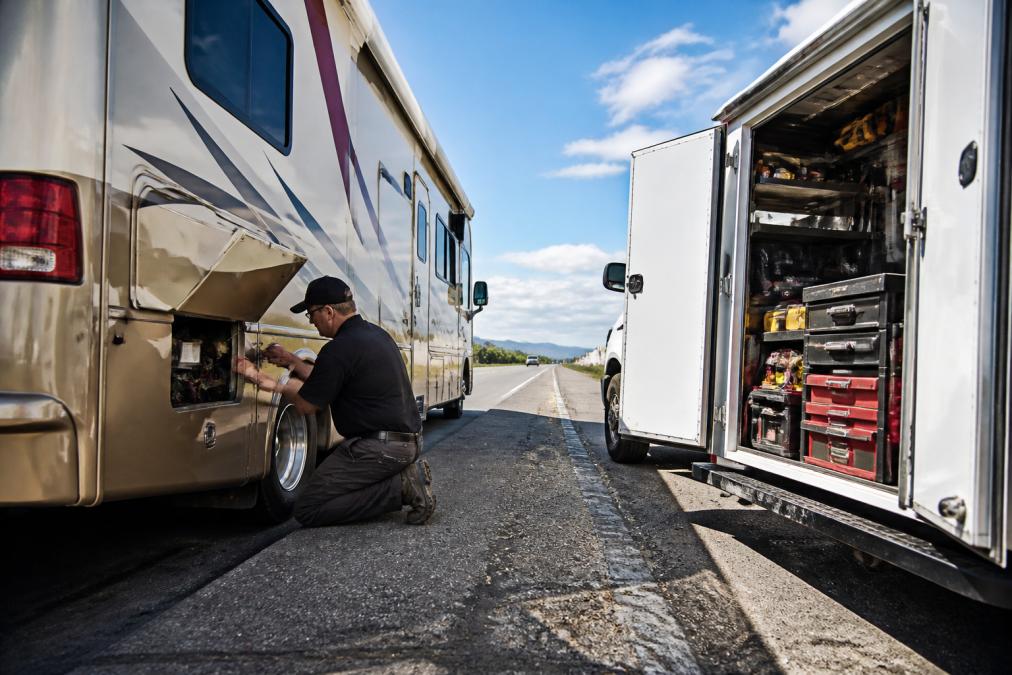 Mobile service truck parked alongside an RV on the shoulder of a highway, technician working on the RV exterior with diagnostic equipment and tools visible, daylight conditions