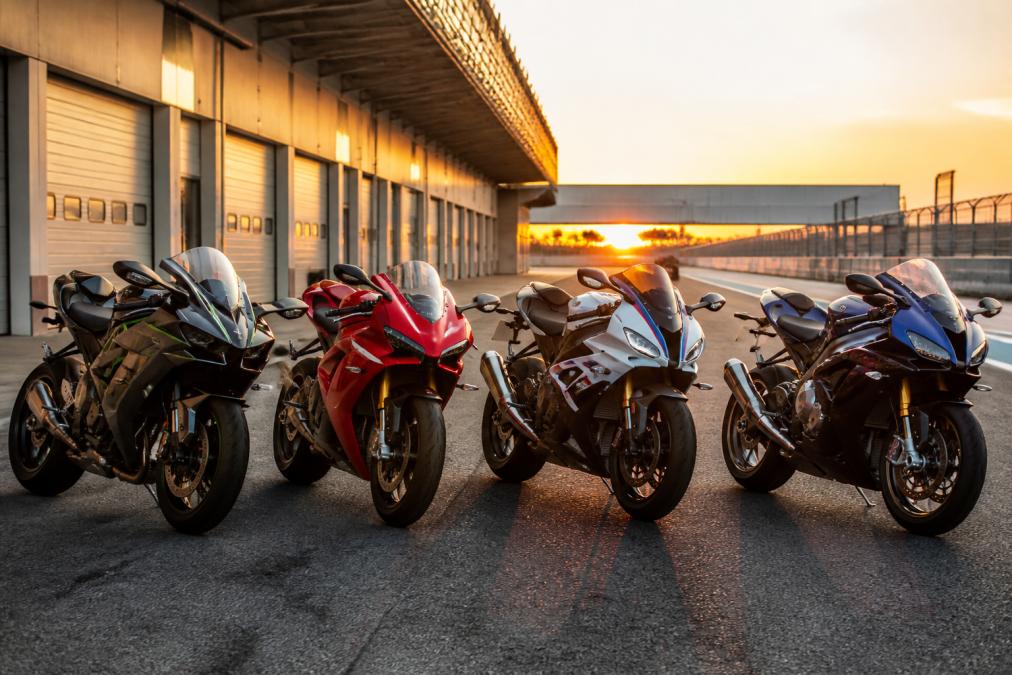 Een rij van vier verschillende sportieve motorfietsen naast elkaar op een circuitparkeerplaats, met op de achtergrond een pitstraat en laagstaande zon die over de motorkappen schijnt