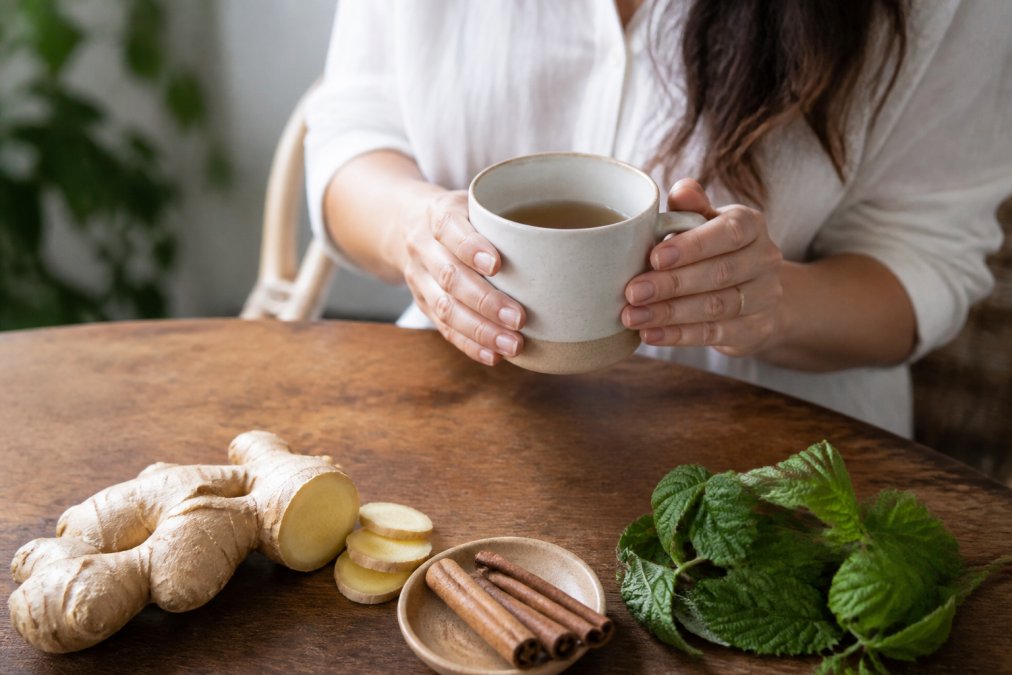 Woman holding a mug of herbal tea, with fresh ginger, cinnamon sticks, and red raspberry leaf visible on a wooden table nearby, natural morning light