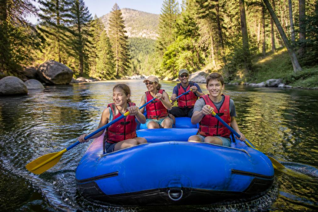 A family with teenagers smiling and paddling a raft on a calm, sun-lit river section surrounded by pine trees and granite boulders, warm afternoon light catching the water surface