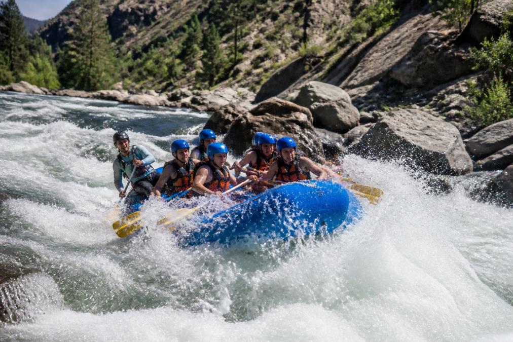 A group of rafters in helmets and life jackets paddling hard through a churning Class IV rapid on a rocky granite river canyon, white water splashing high around both sides of the raft, midday sun lighting the canyon walls