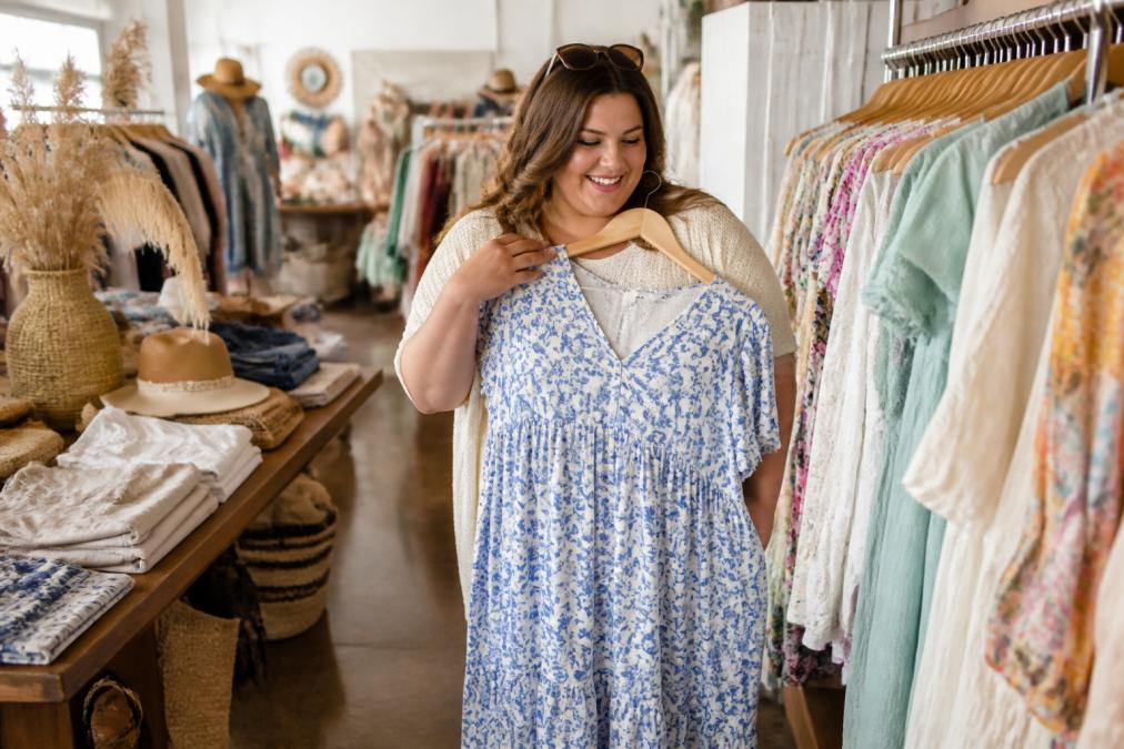A woman browsing a bright, welcoming boutique interior with racks of colorful coastal clothing, holding a dress up to herself with a smile