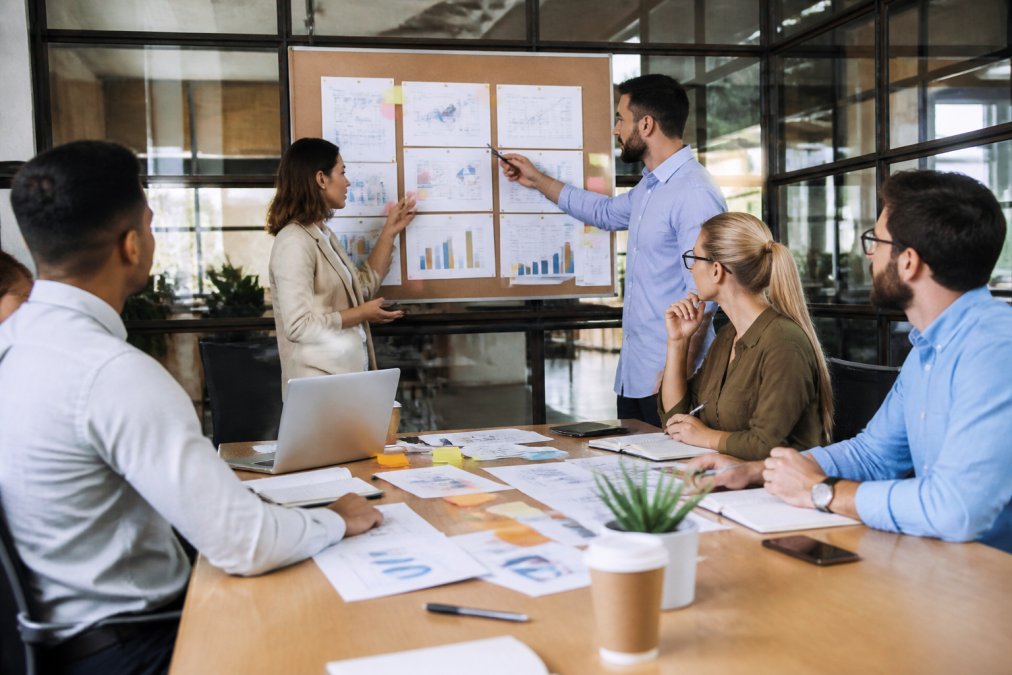 Team in meeting room reviewing performance metrics on large screen with team members pointing at data and discussing strategy
