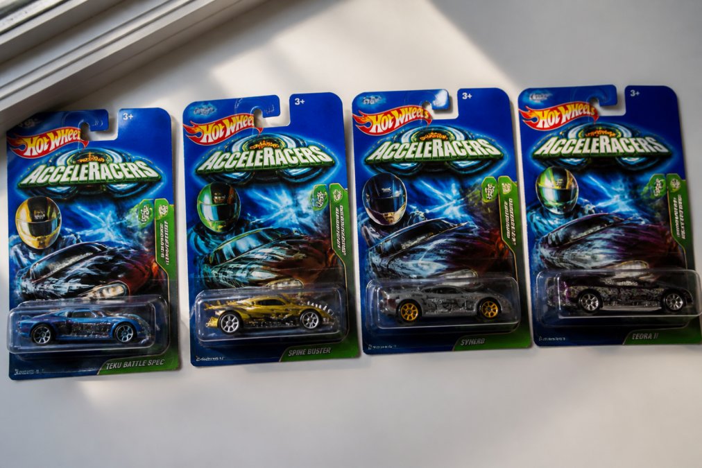 A flat lay of several original carded Hot Wheels AcceleRacers models, including TEKU Battle Spec and Spine Buster, arranged on a clean white surface with natural window lighting showcasing the card art and blister packs