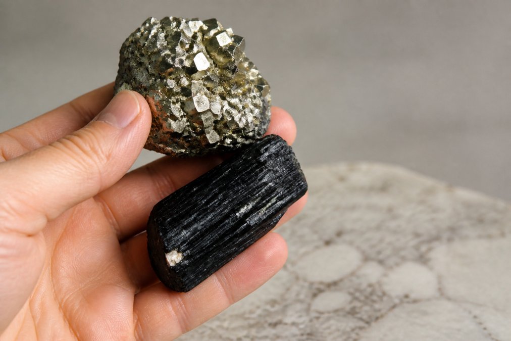 Close-up of a collector's hand carefully examining a raw pyrite specimen and black tourmaline piece against a neutral background, showing the metallic luster and natural texture