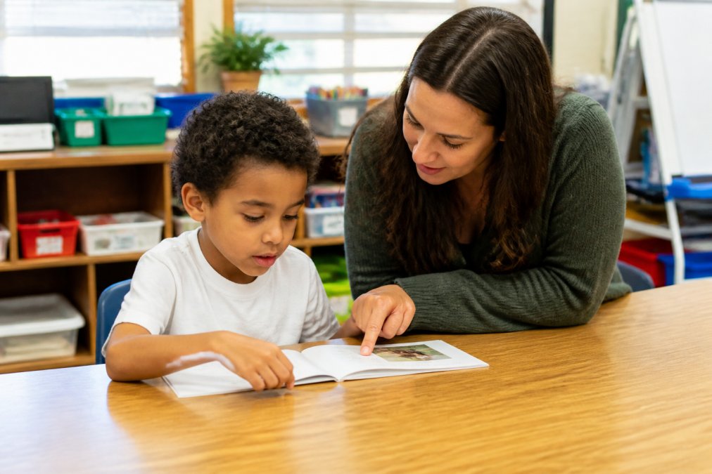 A young student sitting at a classroom table reading a small decodable book, with a teacher kneeling beside them offering guidance, warm natural classroom lighting