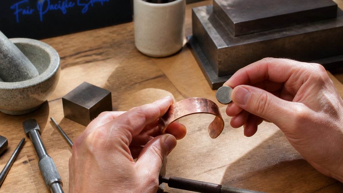 Close-up of hands holding a copper cuff bracelet near a magnet, with natural morning light illuminating the piece's surface patina and warm reddish tones