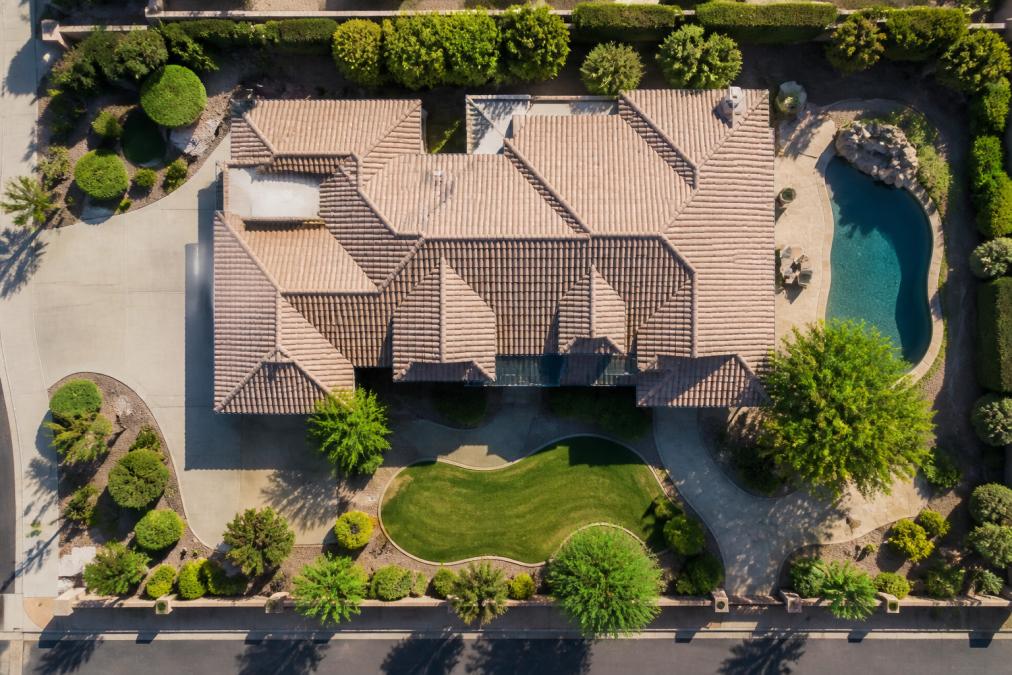 Drone view of residential property from directly overhead showing complete roofline, pool, driveway, and manicured landscaping with clear blue sky and natural sunlight