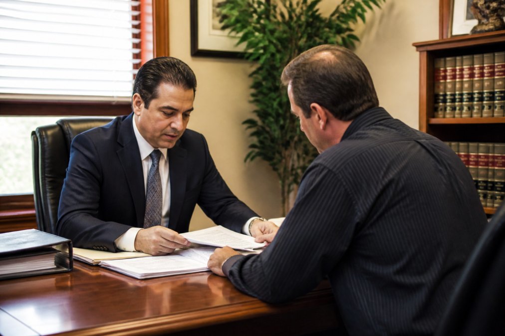 Attorney in a professional office reviewing real estate documents with a client seated across the desk, both focused on papers and a laptop, warm overhead lighting