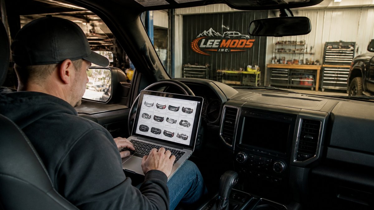 Person inside a truck cab using a laptop to shop for auto parts with tools visible in the garage background